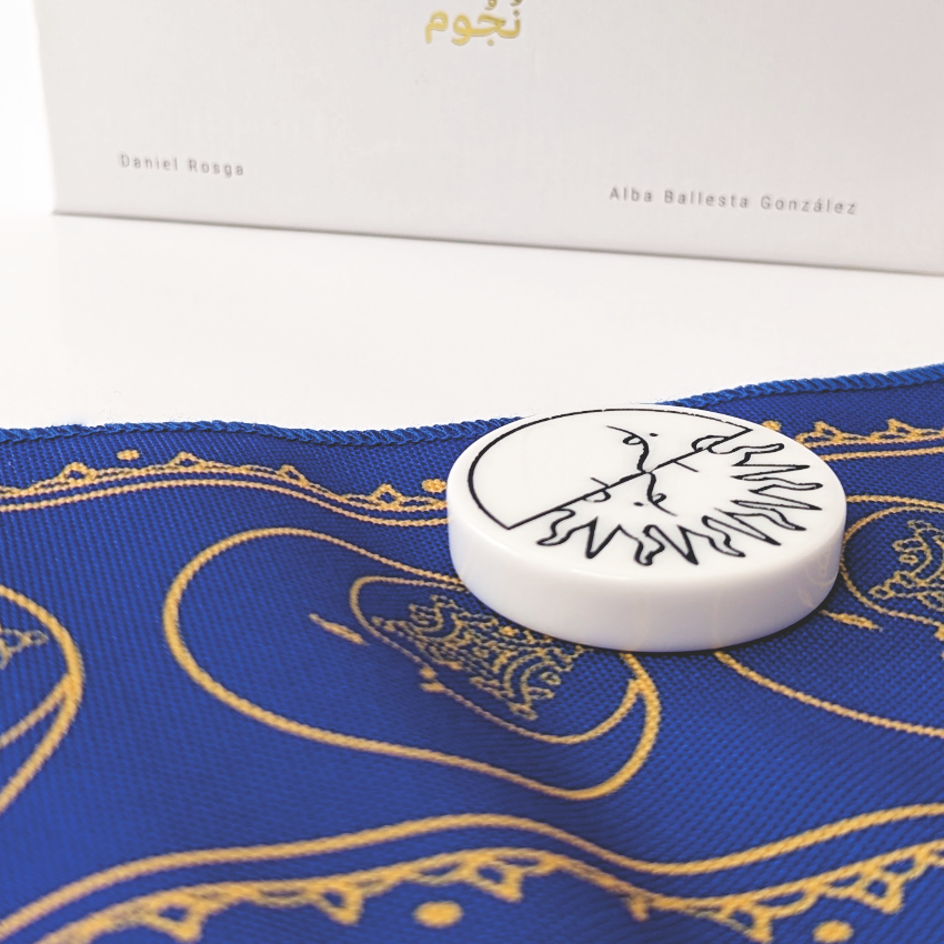Close-up of NUJUM board game components showing a sun and moon token resting on an ornate blue and gold game cloth, with the edge of the game box in the background
